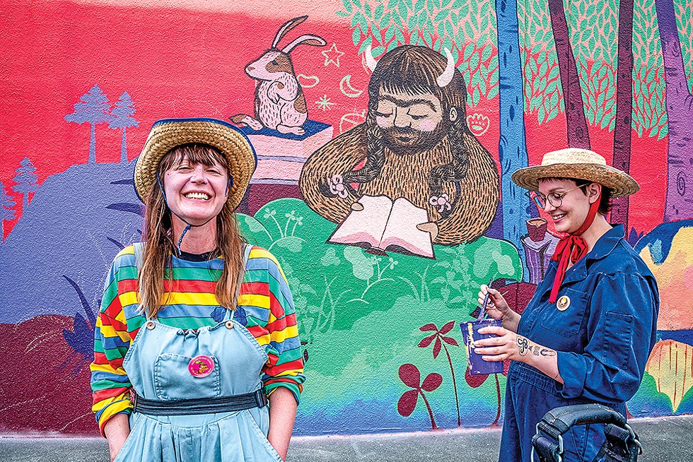 Mural artist Mir de Silva (right) and Violet Crabtree (left), ) explain the backstory of "The Lost Pages" mural design on the side wall of Alder Grove Charter School: "They wanted it to look like a page from a story book, given their love of reading at the school."  She also explained the that she enlisted Crabtree, currently working on her feature film "Wildfur," to help with the mural featuring Wildfur's protagonist, Finnea Wildfur the sasquatch lady. (Team members included Emily Rune, Violet Crabtree, Mir de Silva and Megan Phillips.) - PHOTO BY MARK LARSON