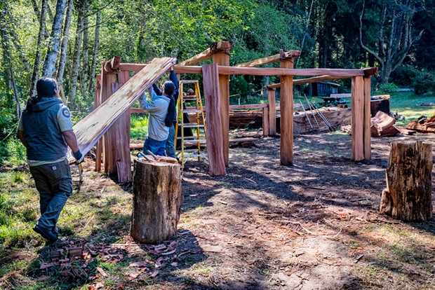 Construction crew members Senior Park Aide Jake Reed (left) and Yurok Trail Crew members Napooi Shorty and Michael Wolf (partially hidden) lift the first of the new roof planks onto the framework of the new women's Dressing House in Sue-meg Village.