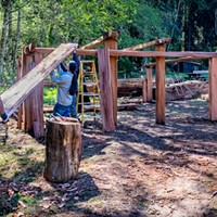 Construction crew members Senior Park Aide Jake Reed (left) and Yurok Trail Crew members Napooi Shorty and Michael Wolf (partially hidden) lift the first of the new roof planks onto the framework of the new women's Dressing House in Sue-meg Village.