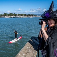 Dancing witch Lisa Wilhelmi Perkins, who chose to stay on land, photographed Ellen McLaughlin, of McKinleyville, on her red-and-white stand-up paddle board and other witch paddlers along the edge of Humboldt Bay with Woodley Island in the background.